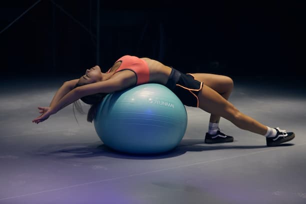 A women is laying her back on a yoga ball in a physio in Tokyo. She is arched in a way that allows the body to stretch. She is located in a dark room with lights focusing on her at a physio in Tokyo.