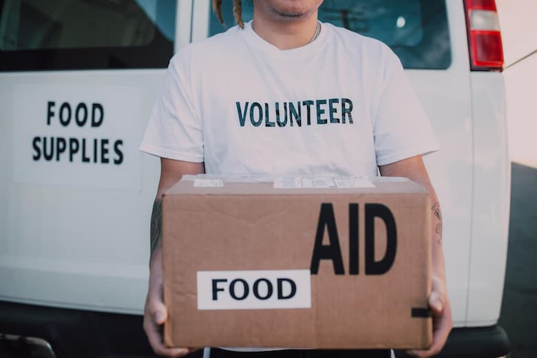 A person is volunteering and he is wearing volunteer shirt holding a cardboard box that reads "food aid". they are standing in front of a white van that says "food supplies" on the back.