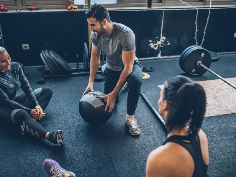 Trainer at the gym showing class how to use equipment properly while people sit on the floor around him and listen