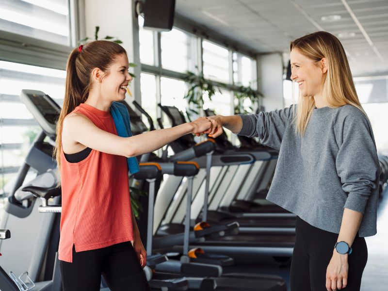 Two women fist bumping at the gym - one wears a red tank top and the other a blue sweatshirt