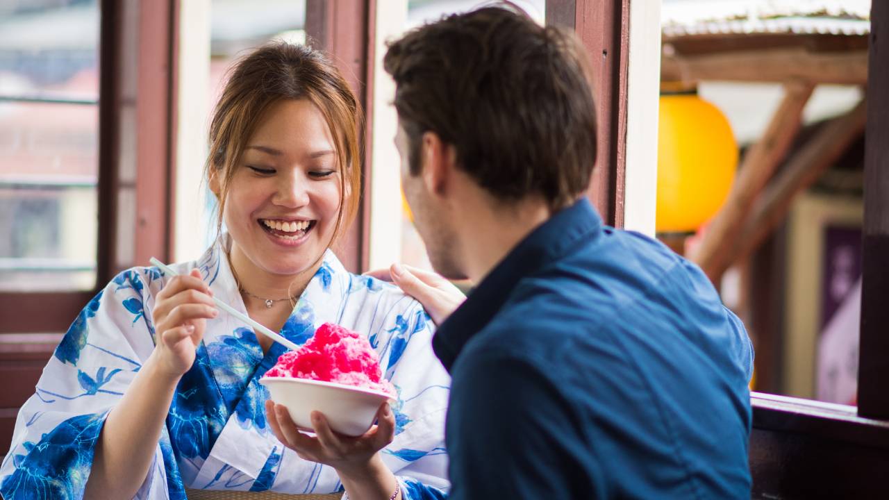 man watches woman smiling while holding spoon over shaved ice