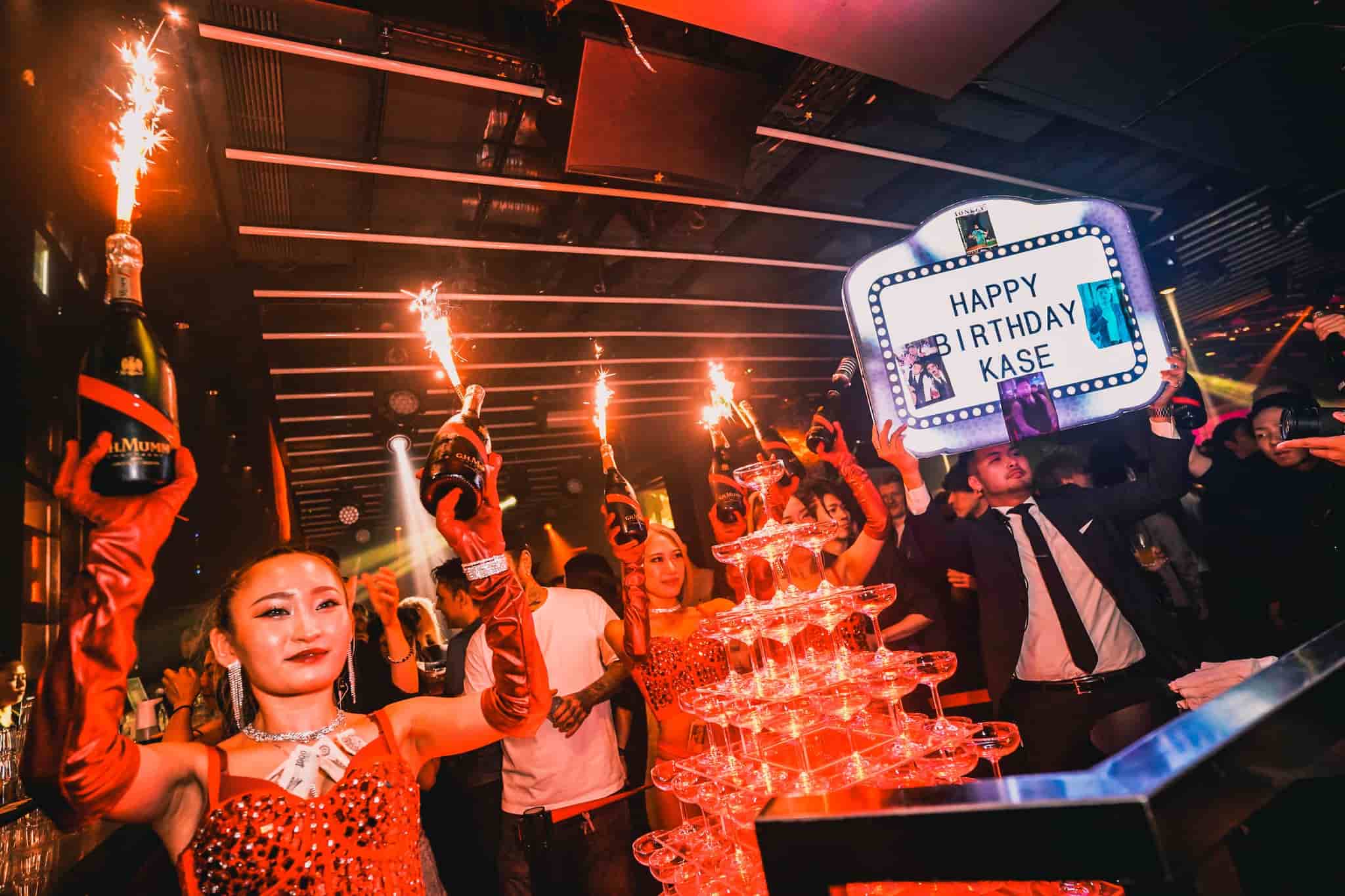 Bottle girls holding sparkling champagne with staff carrying a wish sign for a Shibuya Nightlife birthday bash in a crowded club.
