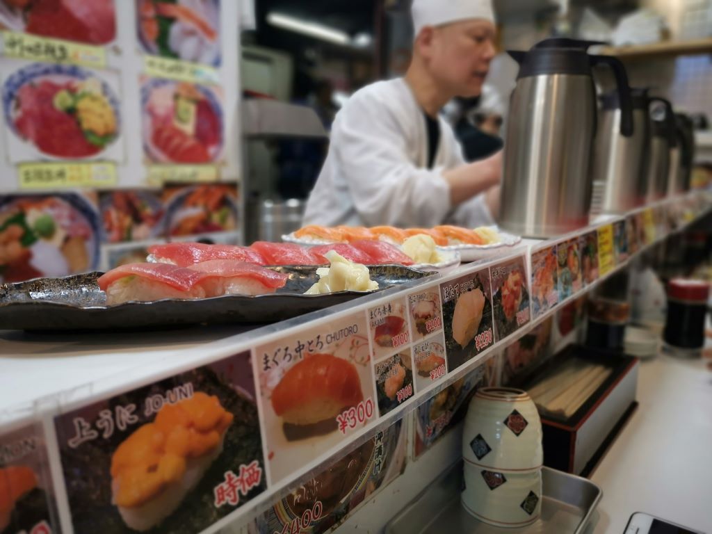 A sushi master is preparing fresh sushi at best sushi restaurant in Tokyo at Fukiji market. In front of him, there are already made sushi dishes, and the restaurant is surrounded by various sushi options with their prices displayed.